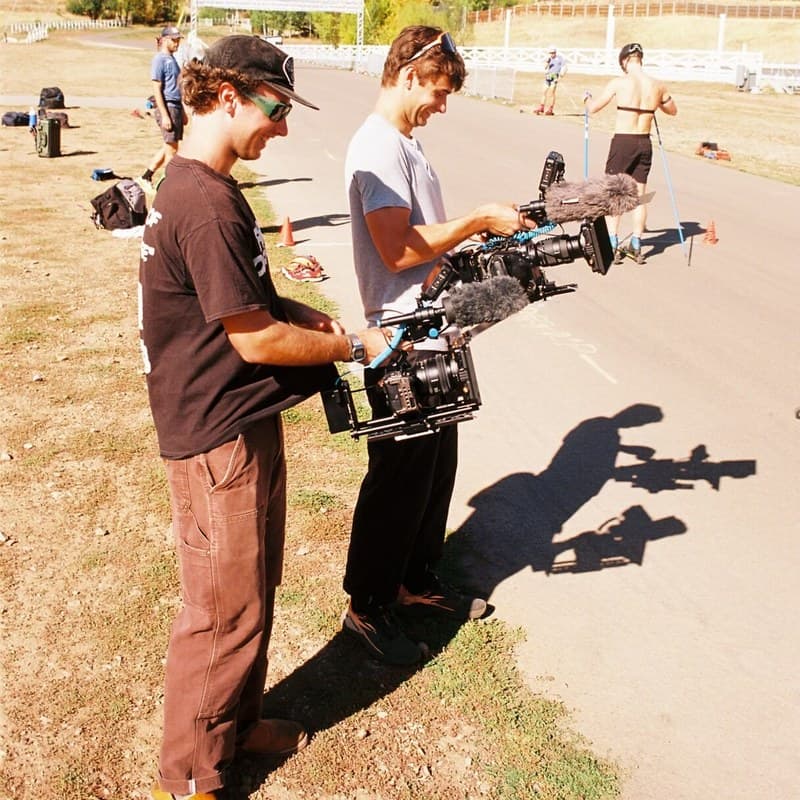 Two filmmakers stand in profile at the edge of a paved roller ski loop, each holding a cinema camera with a microphone mounted on top.