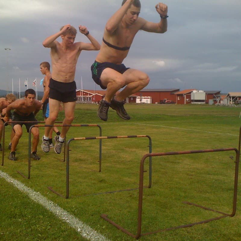 Two runners leap over low hurdles on a grass field during a dryland training session.
