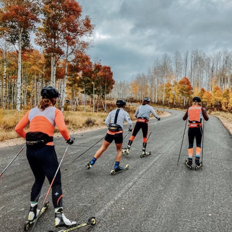 Four rollerskiers skating up a paved road lined with yellow and orange aspens under a cloudy sky