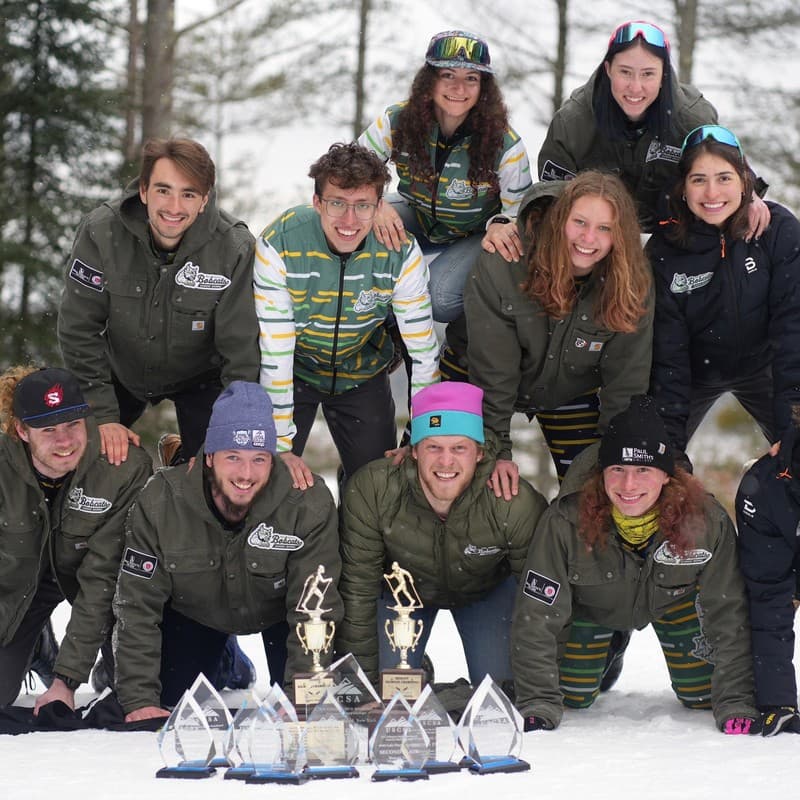 Members of the Paul Smith's College nordic team piled together on snow behind four glass trophies.