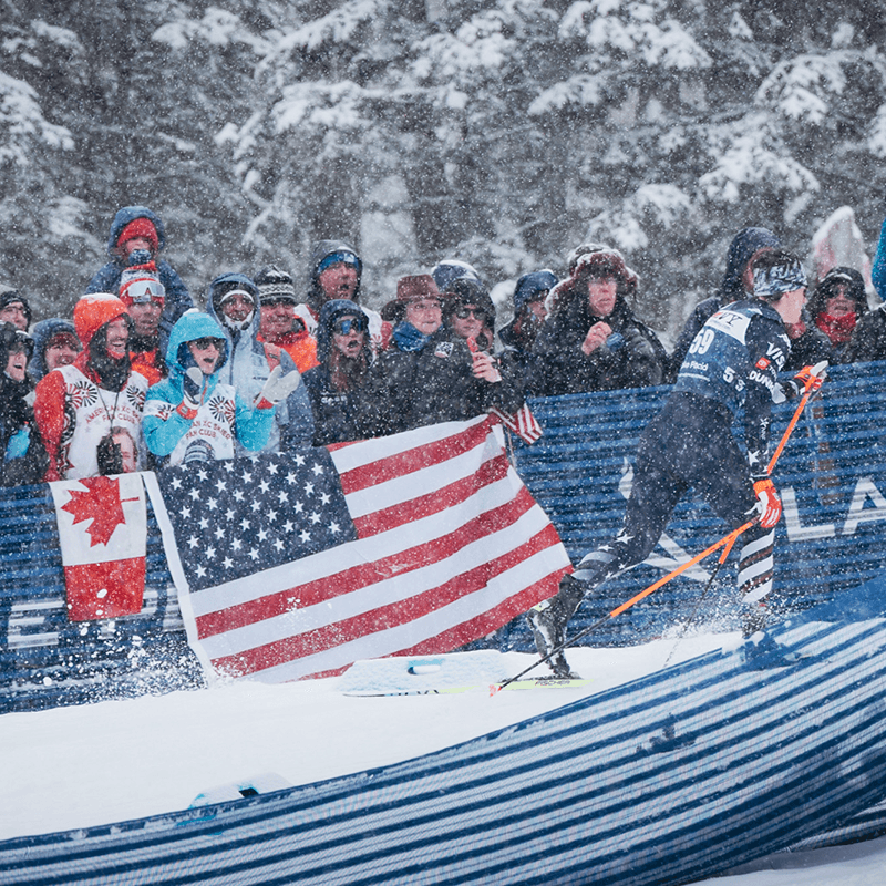 Fans with American and Canadian flags cheering a racer through falling snow in Lake Placid